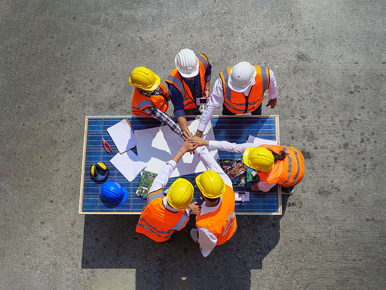 A group of six construction workers in hard hats collaborate around a table covered with plans and materials.